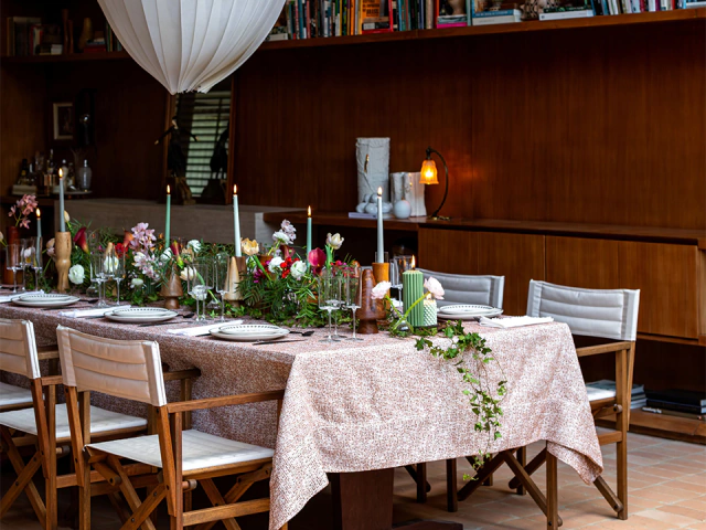 Mesa posta com toalha estampada, flores, velas verdes e brancas e cadeiras de madeira